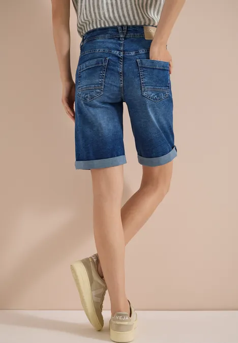 Person von hinten in kniehohen, aufgerollten Jeans-Shorts und beigen Sneakern vor neutralem Hintergrund.