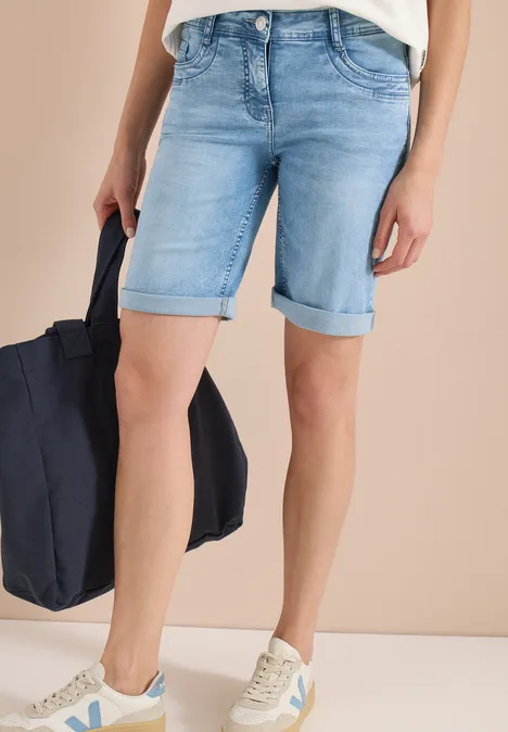 Person trägt hellblaue Jeans-Shorts mit umgeschlagenem Saum, weiße Sneaker und hält eine dunkelblaue Tasche.