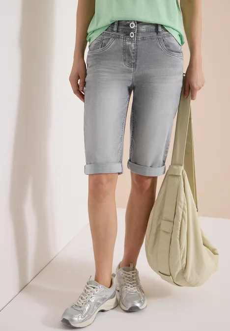 Vrouw in lichtgrijze opgerolde denim shorts, zilverkleurige sneakers, groen topje en grote beige schoudertas.