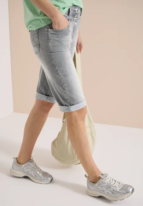 Benen in lichtblauwe, omgeslagen bermudashorts, zilveren sneakers, groen topje en beige canvas tas.