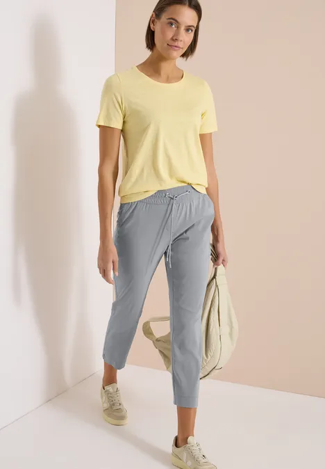 Vrouw in gele T-shirt, grijze 7/8-broek en sneakers, staand met een lichte tas in de hand.