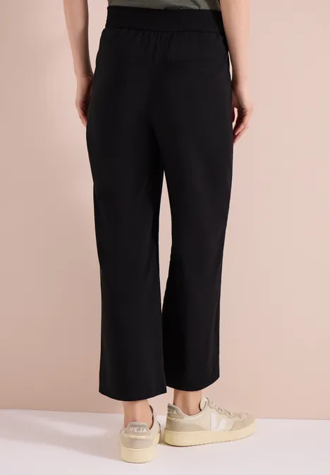 Achteraanzicht van een vrouw in zwarte, wijdvallende, enkellange broek en beige sneakers.