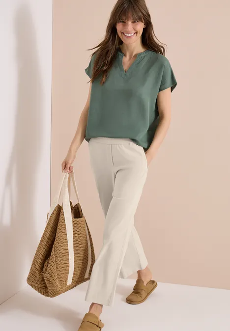 Vrouw met groen topje, witte flared pantalon, rieten tas en sandalen, tegen lichte achtergrond.