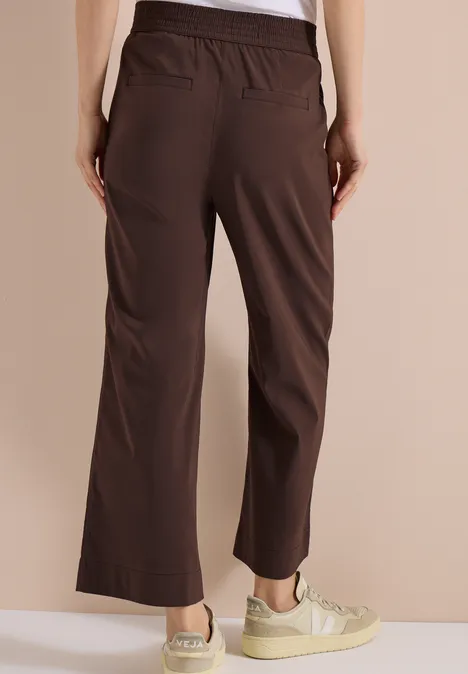 Achteraanzicht van een persoon met losse, bruine broek en lichte sneakers tegen egale achtergrond.