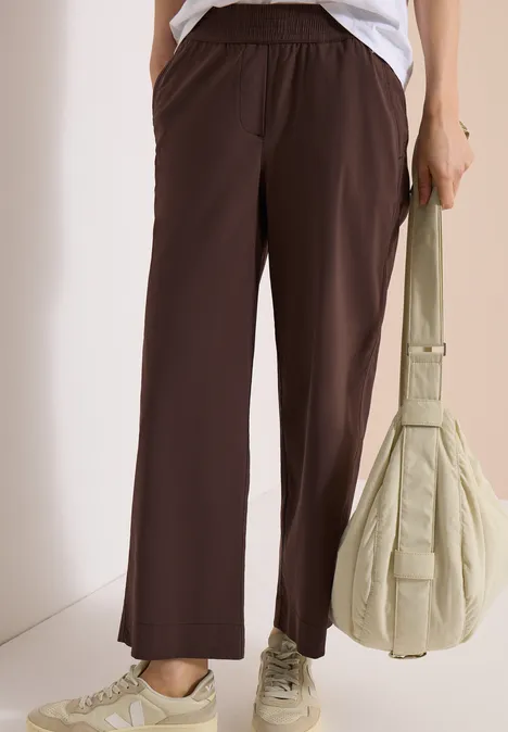 Persoon draagt een losse bruine broek, witte sneakers en heeft een lichte, gewatteerde tas.