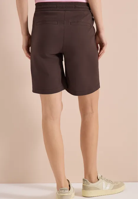 Straight Legs shorts in een Loose Fit brown cacao Achteraanzicht van iemand in donkere bermudashort met zakken, lichte sneakers, beige achtergrond.