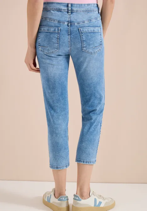 Frau von hinten in hellblauen, hoch sitzenden, knöchellangen Jeans mit weißen Sneakern vor beigem Hintergrund.