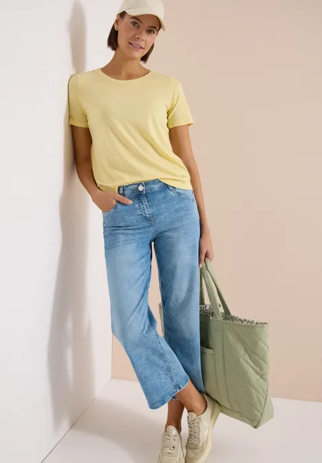 7/8 Wide Leg Jeans im Slim Fit light blue washed Eine Frau trägt ein gelbes T-Shirt, blaue Jeans und hält eine grüne Einkaufstasche.