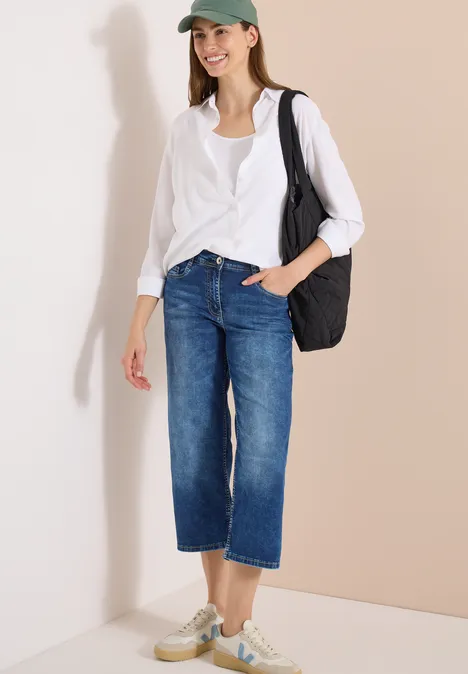 Frau in weißer Bluse, Jeans-Culotte und Sneakern, mit schwarzer Schultertasche vor neutralem Hintergrund.