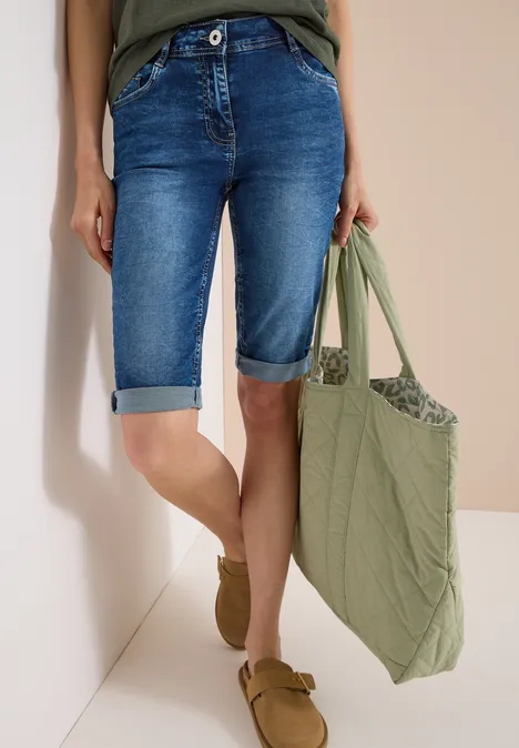 Persoon draagt blauwe denim bermudashort, een kaki shopper en bruine loafers voor een neutrale achtergrond.