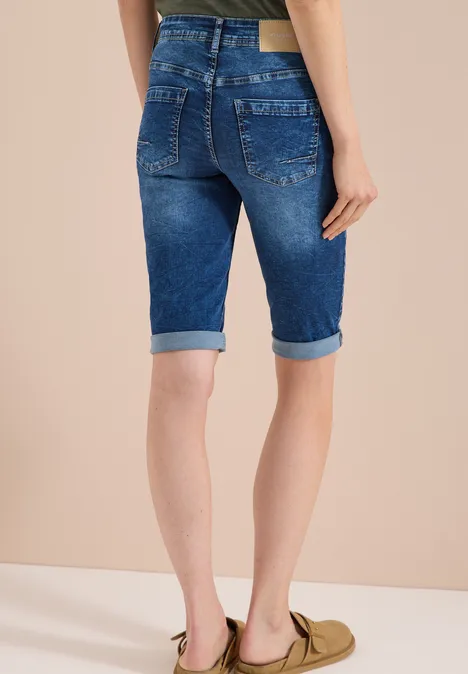 Model draagt blauw omslag-Jeansbermuda en olijfgroene loafers tegen neutrale achtergrond.