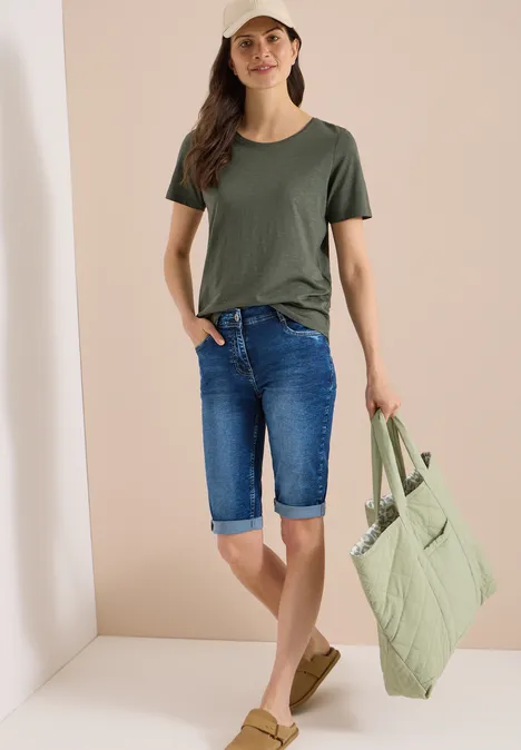Vrouw in olijfgroen T-shirt, denim capri en lichtgroene doorgestikte shopper voor een neutrale achtergrond.