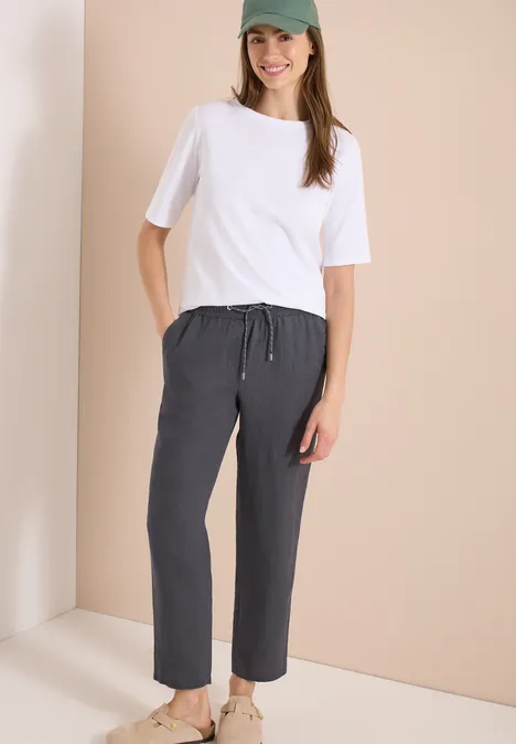 Frau trägt weißes T-Shirt und dunkelgraue, lockere Hose vor beigem Hintergrund.