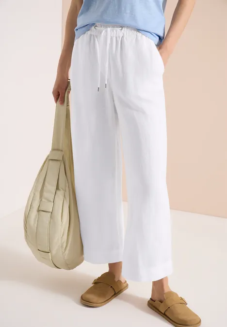 Weiße Leinenhose mit Kordelzug, beige Slipper und große Tasche vor hellem Hintergrund.