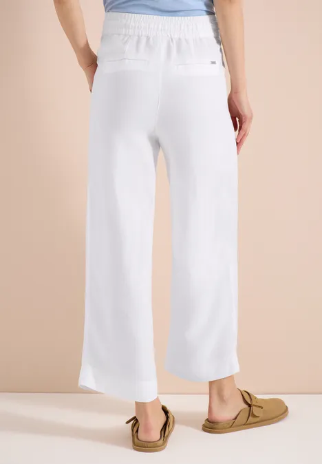 Rückansicht einer Frau in weißen, weit geschnittenen Leinenhosen und Mokassins vor beigem Hintergrund.