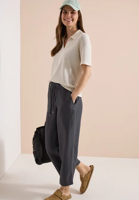 Frau trägt lockeres weißes T-Shirt, weite schwarze Hose, beige Schuhe und hält eine schwarze Tasche.