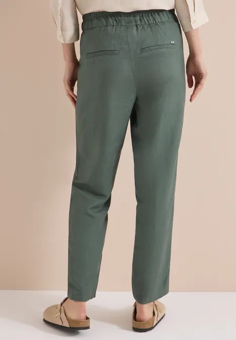 Frau trägt olivegrüne, locker sitzende Hose mit elastischem Bund, aufgesetzten Taschen und flachen Sandalen.