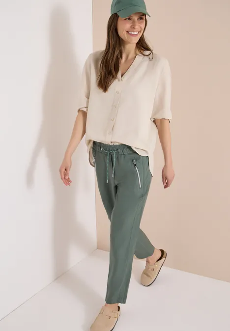 Frau in beiger Leinenbluse, grünen Jogginghosen und Sneakers vor hellem Hintergrund.