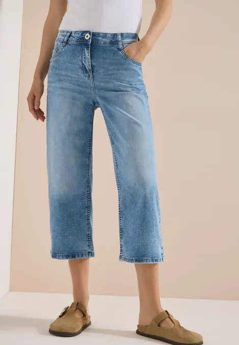 Eine Person trägt hellblaue Culotte-Jeans und beige Slipper vor neutralem Hintergrund.