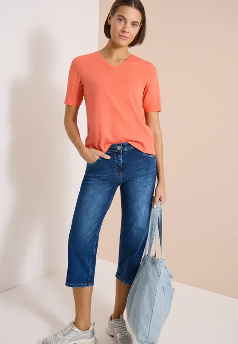 Wide Leg 3/4 Jeans im Loose Fit mid blue used wash Frau in orangefarbenem V-Neck-T-Shirt und blauer Culotte-Jeans mit hellblauer Denim-Tasche vor neutralem Hintergrund.