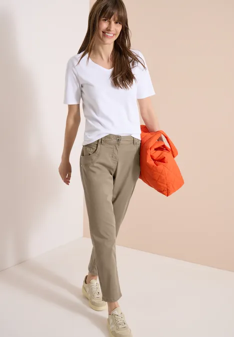 Vrouw in wit T-shirt, kaki broek en witte sneakers, met een oranje tas tegen een lichte achtergrond.