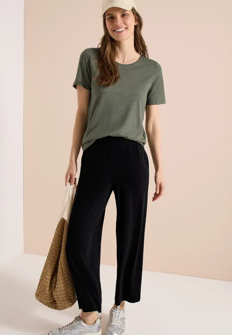 Frau trägt grünes T-Shirt, weite schwarze Hose, weiße Sneaker und Korbtasche vor neutralem Hintergrund.