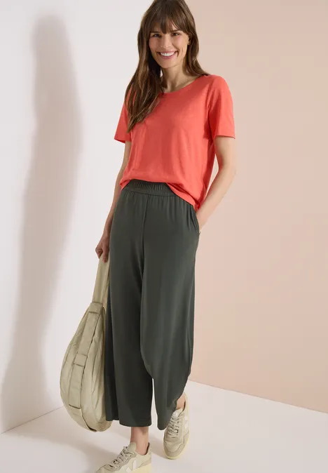 Frau in lachsfarbenem T-Shirt und weiten, dunkelgrünen Balloon-Hosen, trägt eine beige Tragetasche.