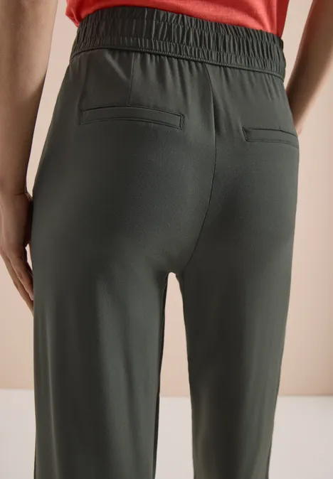 Eine Person von hinten in dunkelgrünen Jogginghosen mit elastischem Bund und Gesäßtaschen vor hellem Hintergrund.