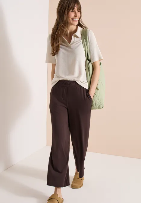Femme souriante en t-shirt blanc, pantalon ample noir et sac vert devant un fond neutre.