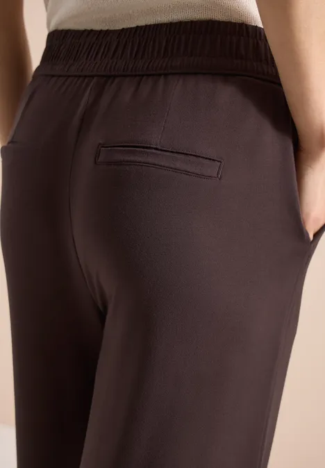 Gros plan d'une personne en pantalon brun foncé à taille élastique avec poches latérales et poche passepoilée.