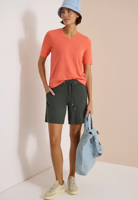 Person trägt korallenfarbenes T-Shirt, dunkelgrüne Shorts, Sneaker und hält eine Jeansjacke.