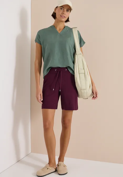 Person trägt grünes Shirt, lila Shorts, Sandalen und Beuteltasche vor hellem Hintergrund.
