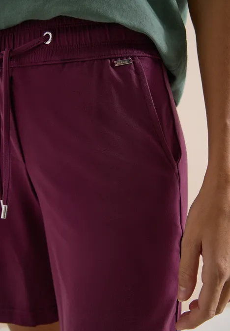 Nahaufnahme einer Person in weinroten Shorts und grünem Shirt, mit Tasche und Kordelzug.