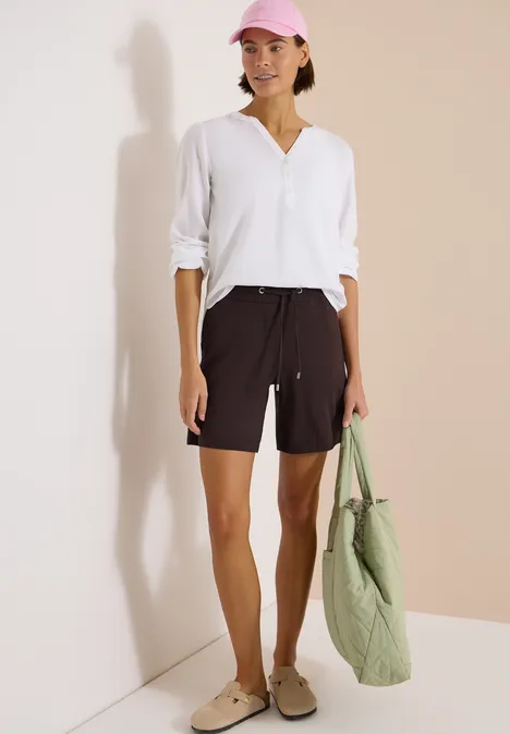 Wide Leg Shorts im Loose Fit brown cacao Frau in weißem Langarmshirt und schwarzen Shorts, beigen Slippers, grüner Stepp-Tasche vor hellem Hintergrund.