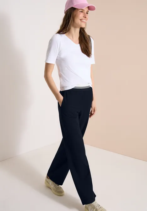 Vrouw in wit T-shirt, zwarte joggingbroek, sneakers en roze pet voor een lichte achtergrond.