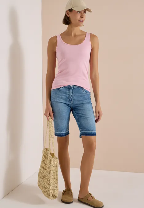 Frau in rosa Tanktop, Jeans-Bermuda, Baseballcap, trägt Strohkorbtasche und bequeme Loafers vor beige Wand.