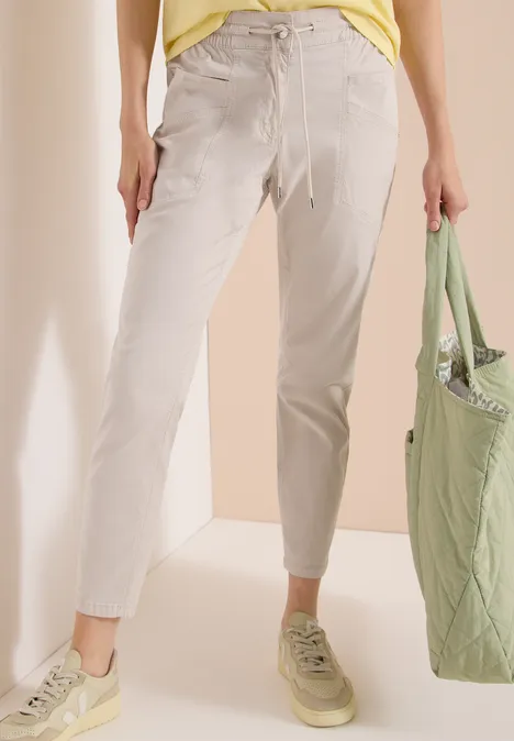 Eine Person trägt helle Stoffhosen, hellbeige Sneaker und hält einen grünen Stoffbeutel.