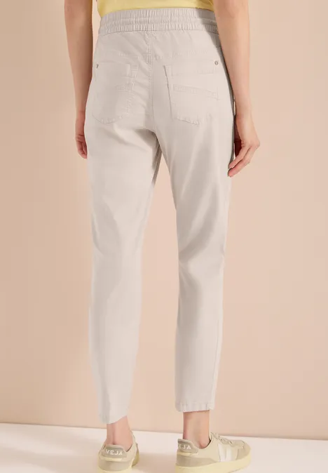 Rückansicht einer Frau in locker sitzender, hellgrauer, hoch taillierter Hose vor beigem Hintergrund.