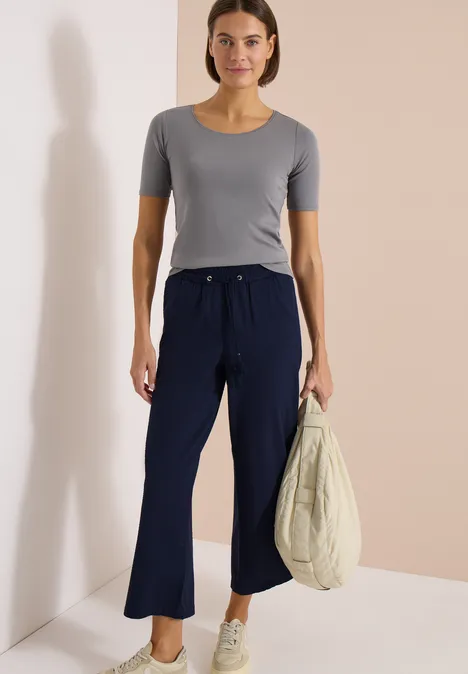 Vrouw met grijs shirt, wijde donkerblauwe broek en tas voor een neutrale achtergrond.