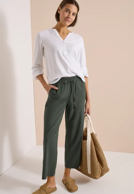 Vrouw in wit overhemd, groene broek en stortas, casual look tegen neutrale achtergrond.