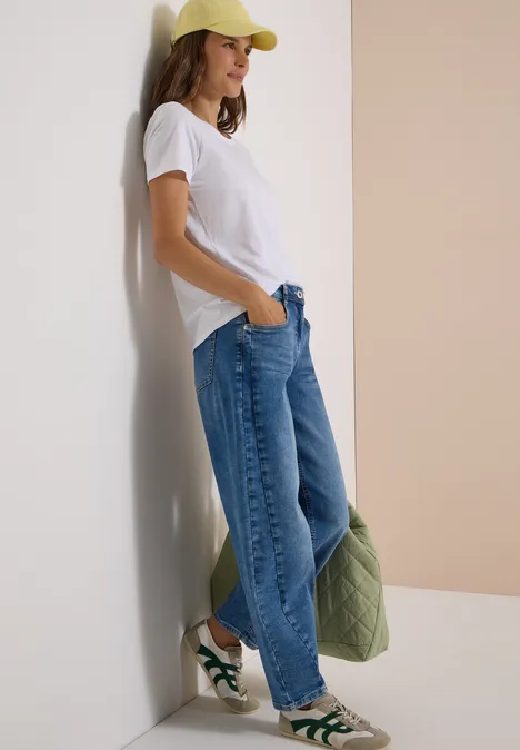High Waist Barrel Leg Jeans im Loose Fit light blue used wash Frau lehnt locker an Wand, trägt weißes Shirt, weite Jeans, gelbe Mütze, grüne Sneaker und hält gesteppte Jacke.