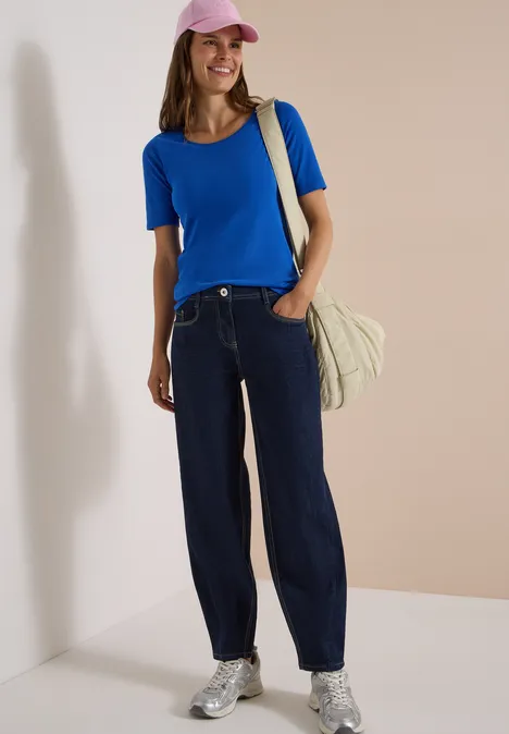 High Waist Barrel Leg Jeans im Loose Fit rinsed wash Frau mit rosa Cap, blauem Shirt, weiten Jeans, silbernen Sneakern und Tasche vor beigem Hintergrund.