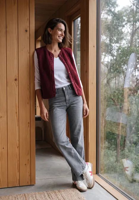 High Waist Wide Leg Jeans im Loose Fit light grey wash Frau in weißem Shirt, grauen Jeans und roter Weste lehnt lässig am Holzrahmen vor einem Fenster.
