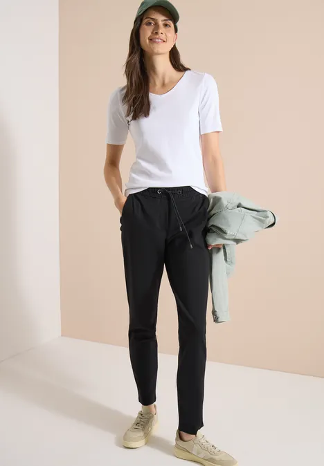 Frau in weißem T-Shirt und schwarzer Hose hält eine Jacke, minimalistische Umgebung, Sneaker an den Füßen.
