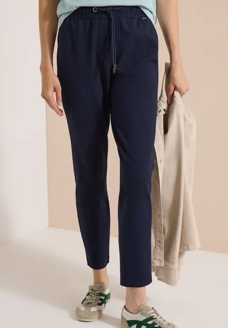 Person trägt dunkelblaue Jogginghose mit seitlichen Streifen, hält beige Jacke und grün-weiße Sneaker.
