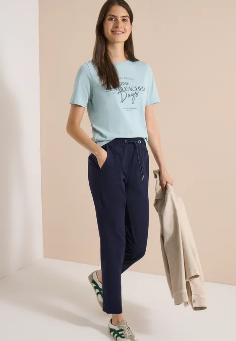 Frau in türkisfarbenem T-Shirt, dunkelblauer Hose und weißen Sneakern, hält beigen Blazer.