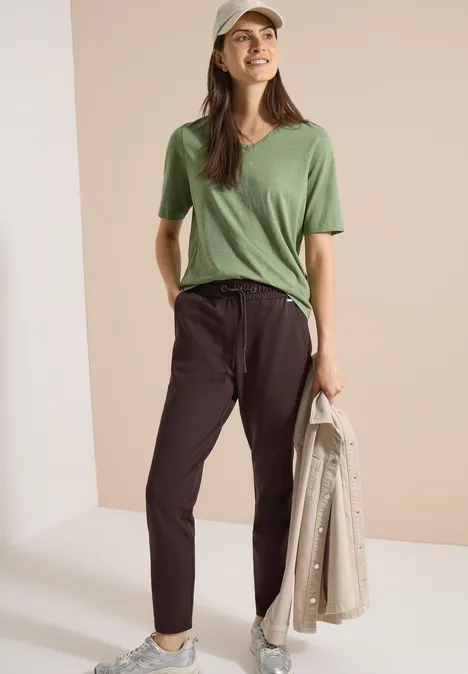 Frau mit Basecap, grünem T-Shirt und schwarzen Stoffhosen hält eine beige Jacke in der Hand.
