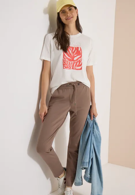 Frau trägt weißes T-Shirt mit rotem Print, beige Hose, hält Jeansjacke vor hellem Hintergrund.