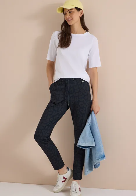 Frau trägt weisses T-Shirt, gemusterte Dunkelhose, weisse Sneaker, gelbe Kappe, hält Jeansjacke.