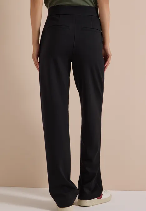 Personne de dos portant un pantalon noir ample et des baskets sur fond neutre.